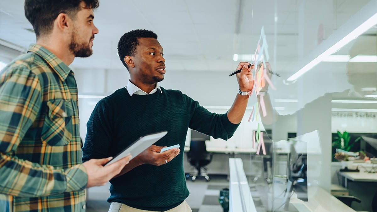 مدیر پروژه در حال نوشتن روی شیشه درحال که همکار مرد در کنارش ایستاده 